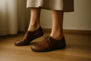 close-up of women's feet, one standing on tiptoes and the other flat, both wearing wide barefoot leather shoes on a wood